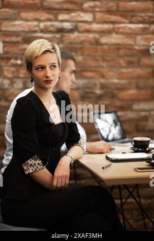 Una giovane donna bionda, vestita con un'elegante giacca nera con polsini con stampa leopardata, si siede attentamente a una riunione di lavoro in un rustico caffè con pareti in mattoni. Foto Stock