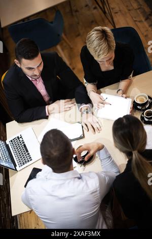 Quattro professionisti collaborano a un tavolo di legno con computer portatili e caffè, mostrando il lavoro di squadra in un ambiente d'ufficio moderno e informale. Foto Stock