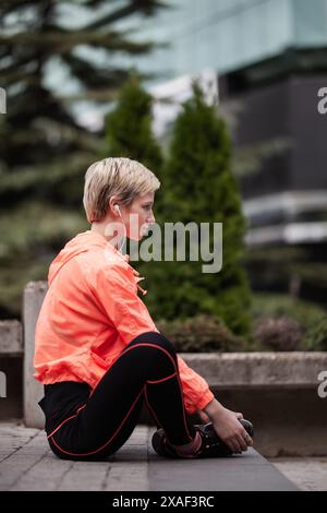 Giovane donna atletica con capelli biondi corti che si prende una pausa durante la sua routine di fitness in un ambiente urbano, riflessivo e concentrato. Foto Stock