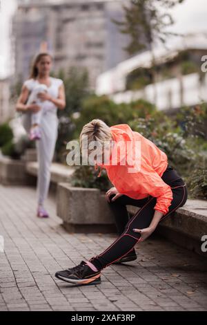Una corsa urbana mattutina catturata con una giovane donna in una vivace giacca arancione che si prepara a fare jogging legando i lacci delle scarpe, mentre un'altra corre con una messa a fuoco morbida Foto Stock