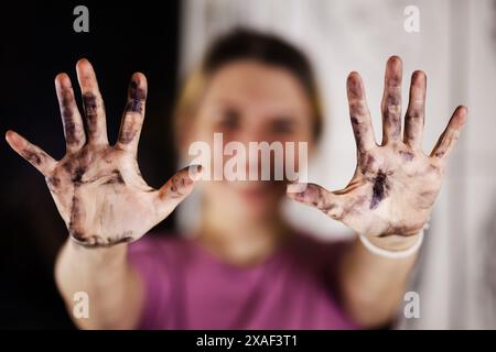 Una gioiosa artista femminile mostra le sue mani ricoperte di vernice verso la fotocamera, mostrando il lato disordinato e divertente della pittura. Foto Stock