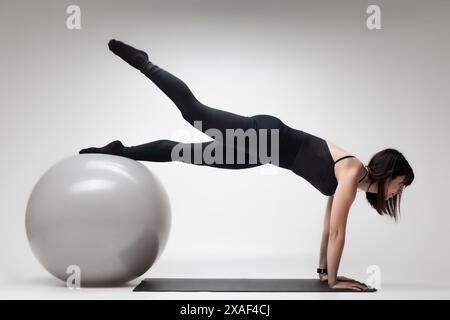 Una giovane donna concentrata si impegna in un esercizio yoga impegnativo utilizzando una palla e un tappetino da fitness in uno studio, mostrando forza ed equilibrio. Foto Stock