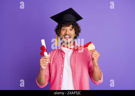 Ritratto di un bel giovane uomo mortarboard diploma medaglia d'oro indossare camicia rosa isolata su sfondo di colore viola Foto Stock