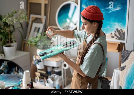 Artista donna incentrata che tiene tele, flacone spray che completa il lavoro finale sulla pittura in resina epossidica Foto Stock