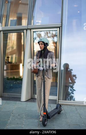 una donna d'affari verticale viene vista uscire da un edificio di uffici con uno scooter elettrico e un casco, mostrando il suo viaggio verso l'esterno nel rispetto dell'ambiente Foto Stock