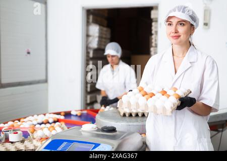 Giovane agricoltrice positiva con cappotto bianco che ordina in base alla taglia e segna le uova di pollo fresche sul nastro trasportatore dalla penna con i polli Foto Stock