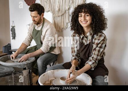 Uomini e donne ceramisti che creano vasi di argilla sulle ruote del vasaio, sorridono e si divertono con attività artistiche in uno studio ben illuminato. Foto Stock