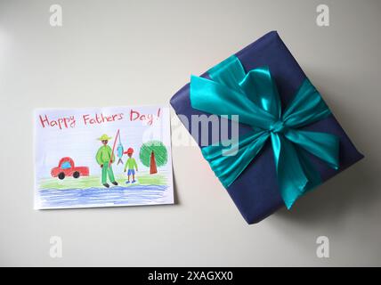 Festa del papà foto di una cartolina disegnata da un bambino. Un regalo di colore blu con un fiocco. Sfondo bianco. La cartolina mostra un padre e un figlio a pescare. AFR Foto Stock