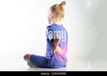 Una ragazza con una tuta viola si siede e le tiene la schiena in basso su uno sfondo bianco. Concetto di dolore e infiammazione nella colonna vertebrale nei bambini. Sprains e st Foto Stock