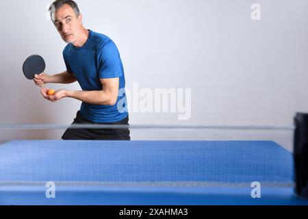 Giocatore di ping pong pronto a servire con una palla arancione in mano dietro un tavolo da gioco blu su uno sfondo bianco isolato. Foto Stock