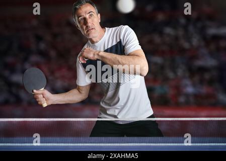 Attento giocatore di ping-pong che riceve una palla bianca dietro un tavolo da gioco blu in una competizione con gli stand sullo sfondo. Foto Stock