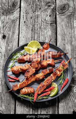 delizioso e succoso kebab di maiale sul piatto con rosmarino, lime e peperoncino rosso secco su un rustico tavolo in legno grigio, vista verticale dall'alto, spazio libero Foto Stock