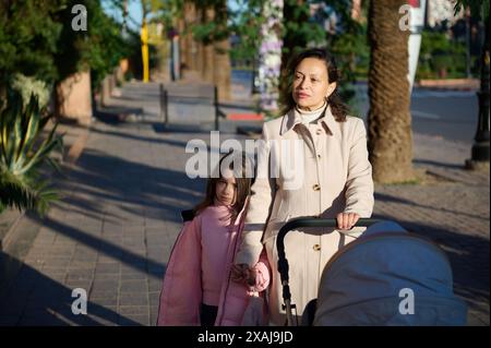 Una madre vestita con un cappotto beige cammina con sua figlia e spinge un passeggino lungo un marciapiede urbano. La figlia in una giacca rosa le regge la m Foto Stock