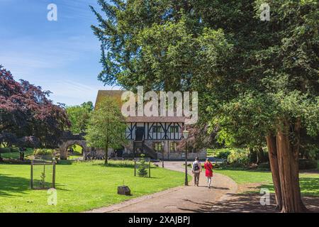 Un sentiero conduce ad un antico edificio in legno situato in un parco comunale. Un grande albero è in primo piano e un paio camminano lungo il sentiero. Foto Stock