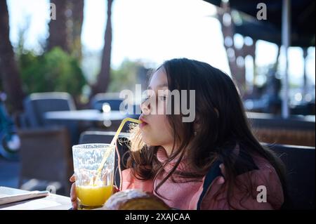 Una giovane ragazza con lunghi capelli castani sorseggia succo d'arancia attraverso una paglia in un caffè all'aperto. Indossa una giacca rosa e si gode un sole rilassante Foto Stock