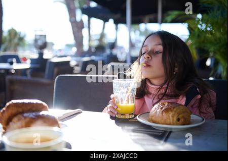 Una ragazza che sorseggia succo d'arancia attraverso una paglia mentre siede in un caffè all'aperto. Si sta godendo una colazione che include un croissant al cioccolato Foto Stock