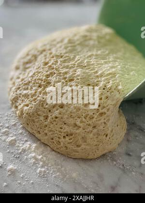 Impasto di pane che scorre dal recipiente, marmo bianco-grigio spolverato di farina Foto Stock