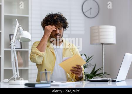 Un giovane sembra stressato mentre legge le cattive notizie ricevute nella posta mentre è seduto alla scrivania del suo ufficio. Foto Stock