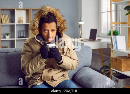 Giovane che si gela a casa in inverno freddo, indossa abiti caldi e beve tè caldo Foto Stock