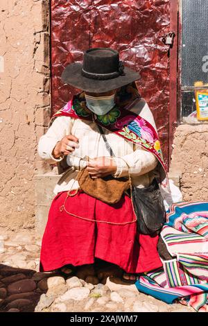 Povera donna adulta indigena artigiana con maschera blu e tipica tessitura ancestrale in tempi di pandemia di covid-19 in una giornata di sole nel mou delle Ande Foto Stock