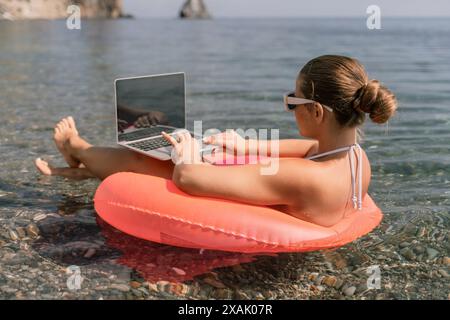 Una donna è seduta su una zattera gonfiabile rosa in acqua, usando un portatile. Concetto di relax e svago, mentre la donna si sta godendo il suo tempo al Foto Stock