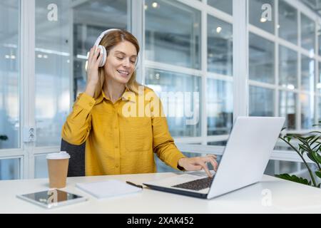 Donna in camicia gialla che utilizza un computer portatile e indossa le cuffie mentre lavora in un ufficio moderno. Ambiente di lavoro mirato e produttivo. Foto Stock