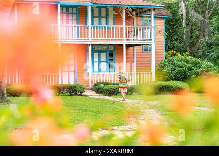 Nord America, Caraibi, isola di Hispaniola, Repubblica Dominicana, Monte Plata, Bayaguana, campo Aventura, donna in abito colorato cammina verso la pittoresca casa di campagna Foto Stock