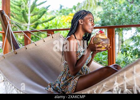 Nord America, Caraibi, isola di Hispaniola, Repubblica Dominicana, penisola di Samana, Sanchez, Jardines la Loma, una bella latina dalla pelle scura si siede su un'amaca bianca e beve da una noce di cocco Foto Stock