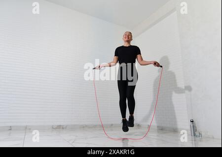 Atleta sportiva che si allena con attrezzature da palestra. Impegnati in una routine di fitness, salta con la corda in uno studio minimalista. Indossa bla Foto Stock