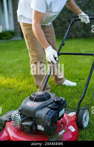 L'uomo irriconoscibile del giardiniere nei guanti di protezione avvia il rasaerba prima di tagliare il prato verde nel suo cortile. Uomo con rasaerba motorizzato ca Foto Stock