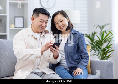 Felice coppia asiatica che si diverte insieme mentre usa uno smartphone sul divano di casa. Sorridente e aderente in un soggiorno accogliente e moderno. Foto Stock