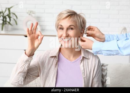 Giovane donna che mette l'apparecchio acustico nell'orecchio di una donna matura mostrando un gesto ok in camera Foto Stock