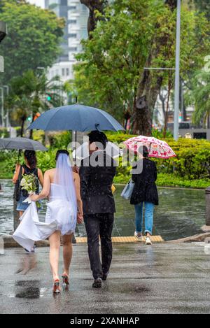 Una giovane sposa e uno sposo attraversa Bras Basah Road a Singapore sotto un ombrello mentre piove verso il centro commerciale Raffles City Foto Stock