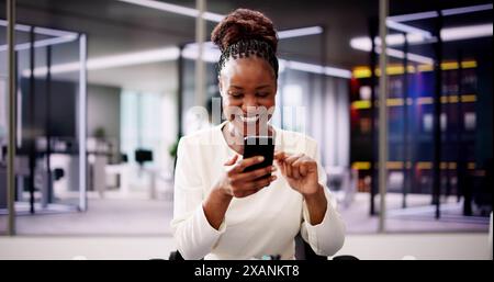 Donna d'affari afroamericana di successo al telefono, festeggia la vittoria alla scrivania con postura felice e smartphone in mano. Foto Stock