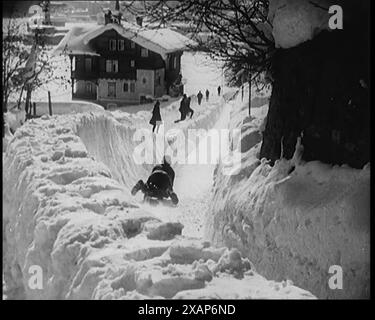 Una donna civile slitta lungo una pista innevata osservata da altri civili con un pittoresco villaggio alpino sullo sfondo, anni '1920 Da "Time to Remember - Teenage Flapper", anni '1920 (Reel 4); un dcumentary sulla vita delle donne negli anni '1920 - grande commento di Joyce Grenfell. Foto Stock