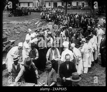 Civili americani che servono pasti per una grande folla in una coda all'aperto, anni '1930 "...l'esercito Bonus [veterani statunitensi che chiedono sostegno al governo durante la depressione] si accamparono alle porte di Washington, chiedendo un trattamento dignitoso... un salario di sussistenza invece di un'esistenza". Da Time to Remember - The Tough Guys, 1930s (Reel 1); un film documentario sulla vita in depressione e sul successo dei gangster in America. Foto Stock