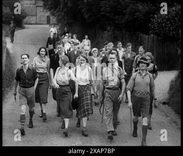 People Hiking Down a Road Towards the camera, 1933. Fu [durante quella brillante estate] che inventarono l'escursionismo. Era come se dopo un secolo di propulsione meccanica, camminare avesse bisogno di un nuovo slogan, di una nuova gimmick per venderlo... l'estate di grandi trekking, l'estate di incontri nei grandi spazi aperti, l'estate di riscoprire l'un l'altro, in pantaloncini corti e camicie". Il massacro di Kinder Scout fu una protesta a Kinder Scout nel Peak District, Derbyshire, che ebbe luogo il 24 aprile 1932. La protesta ha cercato di evidenziare che agli escursionisti è stato negato l'accesso alle aree di campagna aperta wh Foto Stock
