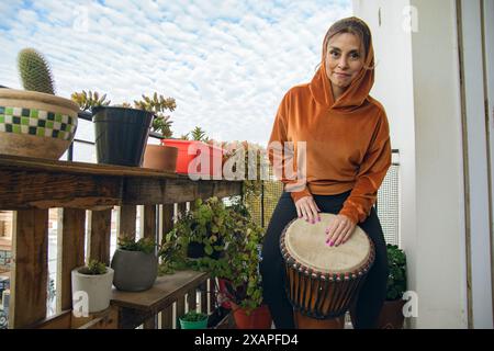 Una donna su un balcone con un tamburo che fa musica africana. Indossa una felpa con cappuccio arancione ed è circondata da piante in vaso. La scena ha una e. Foto Stock