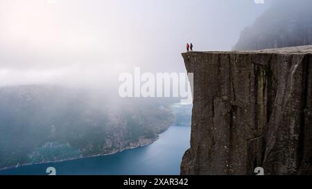 Una vista mozzafiato di due figure in piedi sul bordo di una scogliera torreggiante in Norvegia, che si affaccia su una valle nebbiosa e un fiordo blu profondo. Un paio di uomini e donne che fanno trekking nel Preikestolen, Norvegia Foto Stock