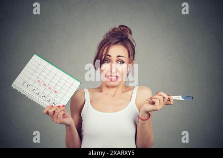Donna con calendario con periodo di assenza contrassegnato e test di gravidanza positivo. Gravidanza indesiderata e ritardo nelle mestruazioni Foto Stock