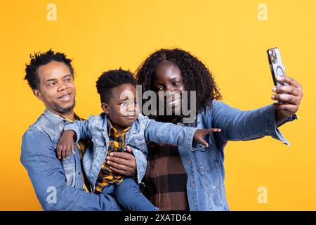 Genitori afroamericani che si fanno selfie con il loro bambino piccolo in studio, posano insieme e fanno ricordi. La mamma e il papà si divertono con il loro bambino, scattano foto al telefono. Foto Stock