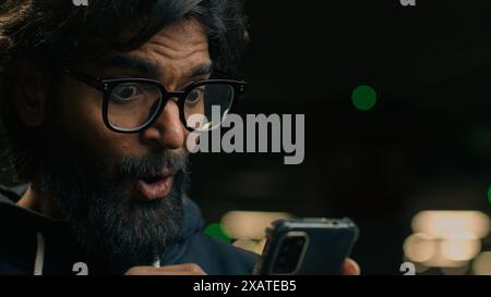 Stupito felice uomo d'affari indiano con la barba in occhiali riceve un messaggio sms leggere buone notizie con lo smartphone nel parcheggio. Vincitore maschio emozionato, Arabian man Foto Stock