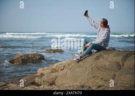 Uomo casual in felpa con cappuccio e jeans seduto sulla roccia in riva al mare, scattando un selfie con uno smartphone. Le spettacolari onde oceaniche e il cielo limpido creano una tranquilla costa Foto Stock