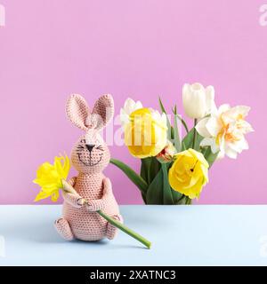 Concetto di primavera, Pasqua, festa della mamma. Coniglietto rosa giocattolo lavorato a maglia con narcisi gialli e bouquet di fiori primaverili di tulipani e narcisi su un tavolo blu e sfondo rosa. Foto Stock