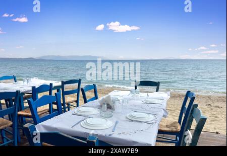 Tradizionale taverna sul mare all'aperto in Grecia. Ristorante Taverna, tavolo vuoto con sedie blu di legno e piatti, spiaggia di sabbia, mare, giorno d'estate. Foto Stock