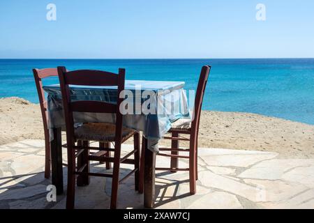 Tradizionale taverna sul mare all'aperto in Grecia. Tavolo vuoto con sedie in legno e tovaglie su un cortile lastricato, spiaggia sabbiosa, mare blu, giorno d'estate. Foto Stock