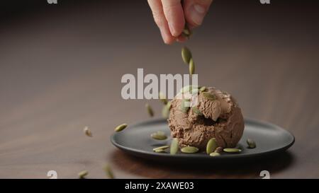 l'uomo ha messo i semi di zucca sopra la palla di gelato al cioccolato su un piattino su un tavolo di legno, foto larga Foto Stock