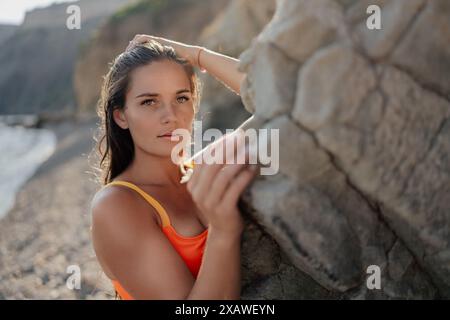 Una donna in una canotta arancione sta potendo per una foto davanti a una roccia. L'immagine ha un'atmosfera da spiaggia e la posa della donna suggerisce che si sta divertendo Foto Stock