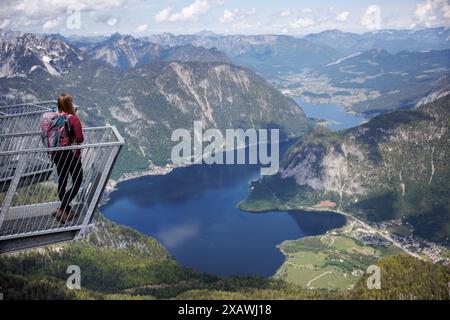 Obertraun, Austria. 20 maggio 2024. Il 20.05.2024, una giovane donna sta con lo zaino da trekking su una delle dita della piattaforma di osservazione a 5 dita sul Krippenstein, una montagna alta 2108 metri sul margine settentrionale dei monti Dachstein nel comune di Obertraun, in alta Austria (Austria), e si affaccia sul panorama montano. Sullo sfondo, il lago Hallstatt con il villaggio di Hallstatt si estende al centro della vista panoramica del paesaggio montano del Salzkammergut dell'Austria superiore. Crediti: Matthias Balk/dpa/Alamy Live News Foto Stock