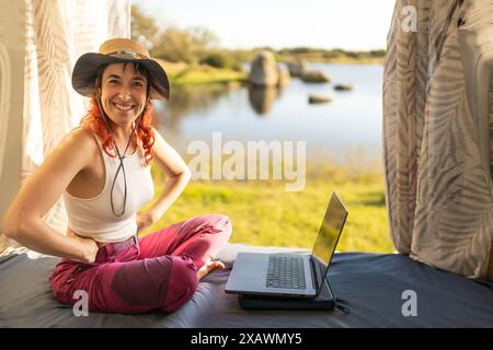 donna che lavora con un notebook in un camper nel mezzo della natura vicino a un lago e a un prato, braccia aperte, con un cappello, al tramonto, nomade digitale, avere un Foto Stock